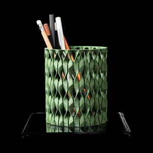 Könnte beinhalten: Ein grüner Stifthalter mit geometrischem Muster und zylindrischer Form. Der Halter ist mit verschiedenen Bleistiften und Stiften in Braun-, Schwarz- und Weißtönen gefüllt. Der Halter steht auf einer schwarzen Oberfläche vor schwarzem Hintergrund.