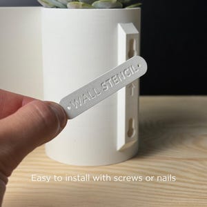 Könnte beinhalten: Ein weißer zylindrischer Pflanztopf mit einer Sukkulente. Ein weißes rechteckiges Schild mit dem geprägten Text WALL STENCIL wird vor den Topf gehalten, der zwei vertikale Schlüssellochschlitze zur Wandmontage aufweist.