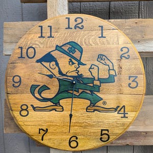 May include: A round wooden clock with a light brown finish. The clock face features the Notre Dame Fighting Irish leprechaun logo in green and navy blue. Black numerals mark the hours. The clock hands are black.