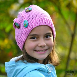 May include: A young girl wearing a pink crocheted hat with colorful buttons. The hat has a unique design with a pointed crown and a wide brim. The girl is smiling and looking at the camera.