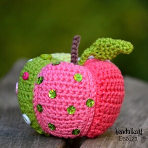 May include: A crocheted apple with a green leaf, pink and green body, and green sequins. The apple is sitting on a wooden surface.