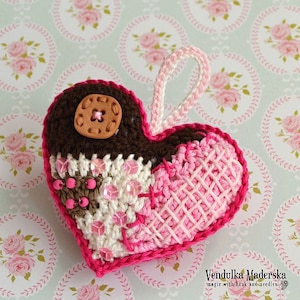 May include: A crocheted heart ornament with a brown button, pink and white crocheted details, and a pink loop for hanging. The heart is on a background of pink and white floral patterned fabric.