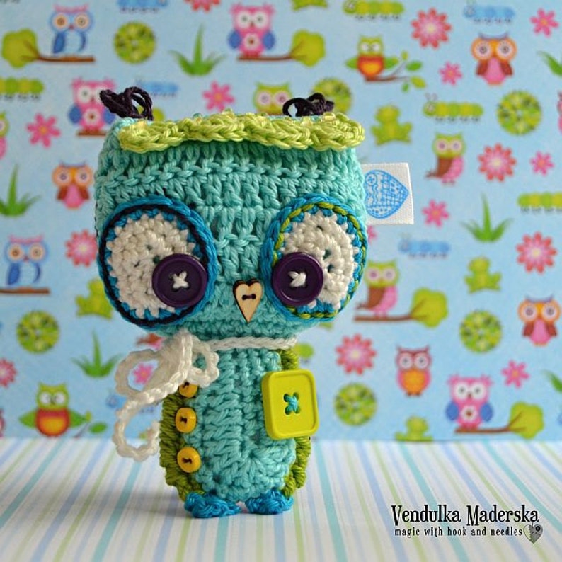 May include: A crocheted teal blue owl with white and purple eyes, green and yellow buttons, and a white scarf. The owl is standing on a blue and white striped surface with a background of a blue and green owl print.