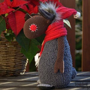 Puede incluir: Figura de alce hecha a mano, tejida a ganchillo, con bufanda y gorro rojos, y una campana en el gorro. El alce tiene una cabeza marr&oacute;n con un dise&ntilde;o cosido en rojo y blanco, un cuerpo gris y una melena gris esponjosa. Sobre una superficie de madera.