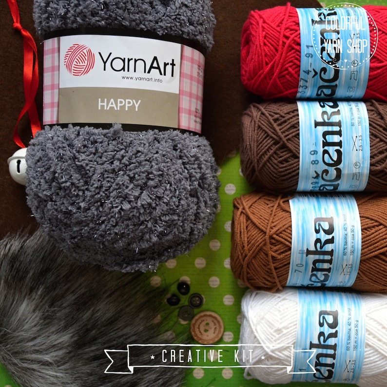 Puede incluir: Una colecci&oacute;n de madejas de hilo en varios colores, incluyendo rojo, marr&oacute;n y blanco, junto con una madeja gris de YarnArt con la etiqueta "HAPPY". La imagen tambi&eacute;n presenta botones, un ribete de piel sint&eacute;tica y el texto "CREATIVE KIT."