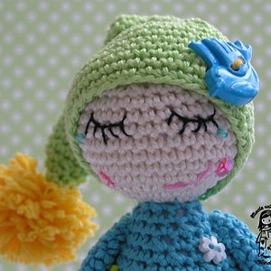 Op de afbeelding: Een gehaakte pop met een groene muts, blauwe ogen en een gele pompon. De pop draagt een blauwe gehaakte jurk en heeft een blauwe bloem op haar borst. De pop is gemaakt met liefde.