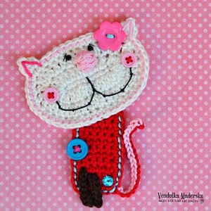 May include: White crocheted cat bookmark with pink flower, red body, and blue buttons. The cat has a black embroidered smile and pink nose.