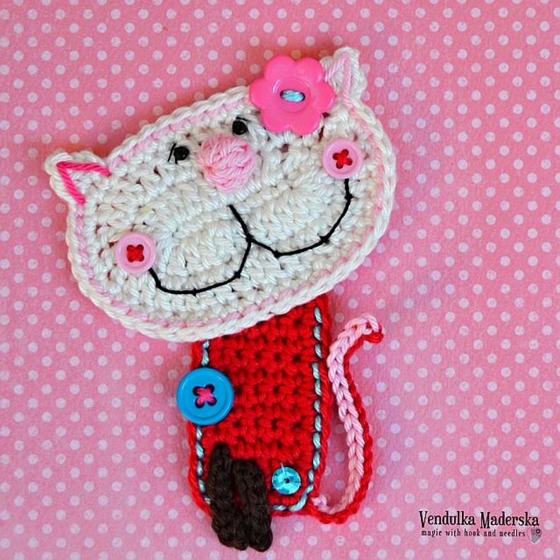 May include: White crocheted cat bookmark with pink flower, red body, and blue buttons. The cat has a black embroidered smile and pink nose.
