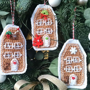 May include: Three crocheted gingerbread house ornaments hanging on a green Christmas tree. The ornaments are brown and white with white trim and various decorations, including a reindeer, a Christmas tree, and a snowflake.