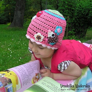 May include: A young girl wearing a pink crocheted hat with colorful flowers and a bird applique. The hat has a scalloped edge and is decorated with buttons. The girl is reading a book and is lying on a blanket.