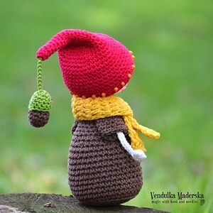 May include: A crocheted gnome doll with a red hat, yellow scarf, and brown body. The gnome is holding a green acorn.