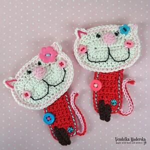 May include: Two crocheted cat bookmarks with red, white, and pink yarn. The cats have black eyes, pink noses, and white teeth. Each cat has a button on its chest and a flower on its head. The bookmarks are on a pink polka dot background.