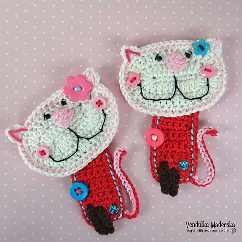 May include: Two crocheted cat bookmarks with red, white, and pink yarn. The cats have black eyes, pink noses, and white teeth. Each cat has a button on its chest and a flower on its head. The bookmarks are on a pink polka dot background.