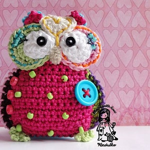 May include: A colourful crocheted owl with large black eyes, a pink body, and a blue button. The owl is decorated with green, yellow, and orange yarn and beads. The background is a pink pattern of hearts.