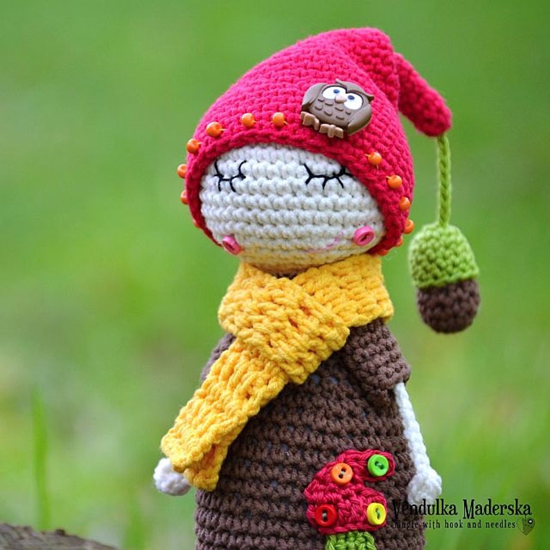 May include: Crocheted doll wearing a red hat with an owl button, a yellow scarf, and a brown dress with buttons. The doll is holding a green acorn.