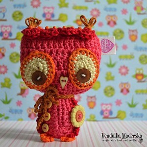 May include: A crocheted pink owl with orange and yellow accents. The owl has large brown button eyes and a small heart-shaped button on its chest. The owl is wearing a brown scarf and has a yellow flower on its wing.