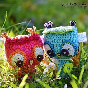 May include: Two crocheted owl toys, one pink and orange and one blue and green, are sitting in a bed of green grass. The owls have large button eyes and are handmade.