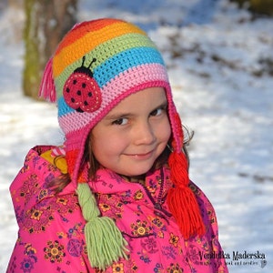 May include: A young girl wearing a colorful crocheted hat with a ladybug applique and red and green tassels. The hat is striped with pink, blue, green, orange, and yellow yarn. The girl is wearing a pink jacket with a floral pattern.