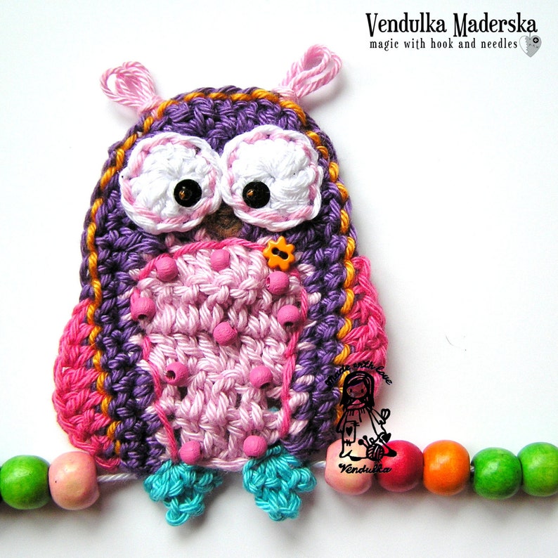 Peut inclure: Un hibou crochet&eacute; &agrave; la main, aux couleurs violettes, roses et blanches, avec des yeux en boutons noirs et une petite fleur orange. Le hibou est attach&eacute; &agrave; une guirlande de perles en bois color&eacute;es. Le texte "Vendulka Maderska" est visible.