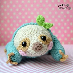 Puede incluir: Un juguete de peluche de perezoso tejido a crochet con un cuerpo azul claro, ojos marrones y una nariz rosa. El perezoso lleva una hoja verde en la cabeza.