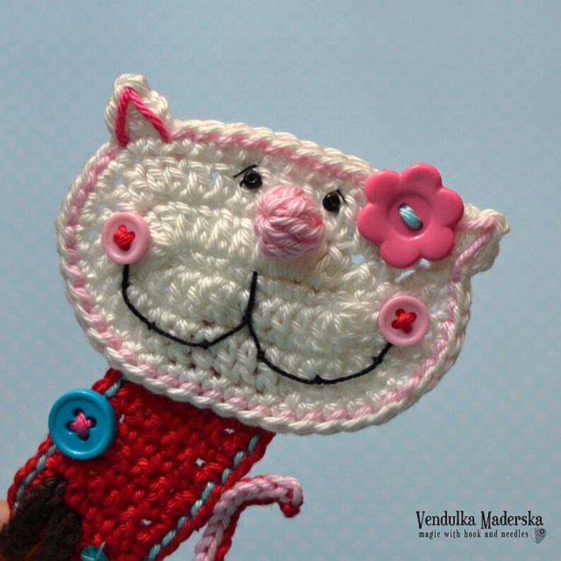 May include: White crocheted cat bookmark with pink and red buttons, a pink flower with a blue button, and black embroidered details. Vendulka Maderska magic with hook and needles.