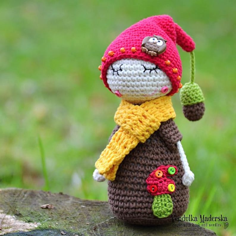 May include: A crocheted doll wearing a red hat with an owl button, a yellow scarf, and a brown dress with a mushroom applique. The doll is sitting on a tree stump.