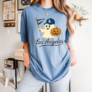 Puede incluir: Camiseta azul claro con un fantasma de dibujos animados con gorra de béisbol y una bandera de los Dodgers, con una calabaza y la inscripción "Los Angeles". La camiseta es de estilo casual.