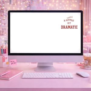 May include: A desktop computer with a white screen sits on a pink desk. The screen displays the text "Lotta A Little Bit Dramatic" in red. A keyboard, mouse, and notebook are also on the desk.