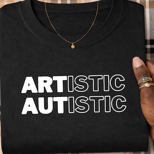 May include: Black crew neck sweatshirt with the words "ARTISTIC AUTISTIC" in white block letters. A gold necklace with a small pendant is visible. The sweatshirt is on a plaid surface.