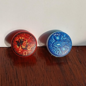Op de afbeelding: Twee ronde, decoratieve pluggen. Eén is rood met een vurig ontwerp en een Omega-symbool. De andere is blauw met een wervelend, aquatisch patroon. Beide hebben een lichtgekleurde rand.