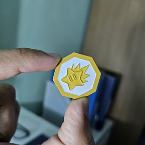 May include: An octagonal coin in yellow and white, featuring a starburst design and the number "00". The coin is held between fingers, with a blurred background. The coin's design is reminiscent of a game token.