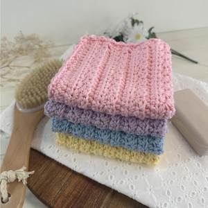 May include: A stack of four handmade crochet washcloths in pastel colors: pink, lavender, blue, and yellow. The washcloths are neatly folded and stacked on a white, eyelet-embroidered cloth. A wooden handled brush and a bar of soap are also visible.