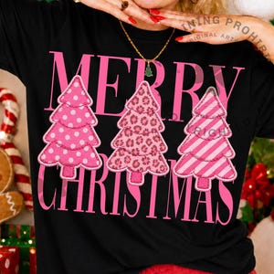 May include: A black short-sleeved t-shirt featuring the pink text "MERRY CHRISTMAS" and three stylized pink Christmas tree graphics. The trees display distinct patterns: white polka dots, a leopard print, and diagonal stripes.