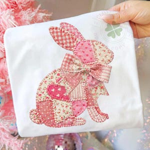 May include: White t-shirt featuring a patchwork bunny design in shades of pink. The bunny has a large bow and is made of various patterned fabrics, including gingham and floral prints. The design is centered on the shirt, creating a charming and festive look.