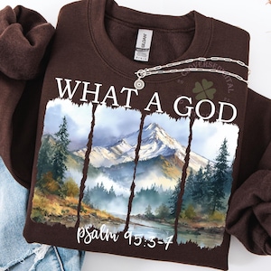 Può includere: Una felpa marrone con la scritta "WHAT A GOD" e una scena di montagna ad acquerello. Il testo "psalm 95:3-7" è sotto l'immagine. Una collana d'argento è sulla felpa. Jeans blu chiaro sono accanto alla felpa.