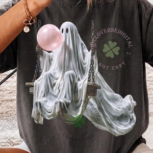 May include: A charcoal grey short-sleeve t-shirt featuring a graphic of a white ghost sitting on a swing and blowing a pink bubble. The design includes a circular emblem with a green four-leaf clover and the text CLOVERSEDIGITAL NOT COPY.