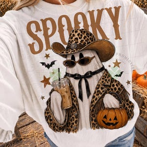 May include: A white crewneck sweatshirt featuring the brown text SPOOKY above a graphic of a ghost wearing a leopard print cowboy hat, sunglasses, and a matching jacket. The ghost holds an iced coffee and a pumpkin bucket with a black bow.