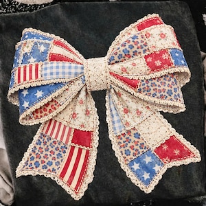 May include: A large, decorative bow with a patchwork design featuring red, white, and blue fabrics. The bow's design incorporates stars, stripes, and floral patterns, with a lace trim. The bow is likely handmade and suitable for patriotic or Americana-themed decor.