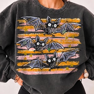 Op de afbeelding: Een antracietgrijze sweater met ronde hals en een grafische print van drie zwarte vleermuizen met grote witte ogen. De vleermuizen zijn geplaatst over horizontale, verweerde penseelstreekstrepen in oranje en roze, geaccentueerd met kleine gele sterren.