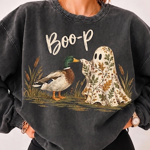 May include: A charcoal grey mineral-washed crewneck sweatshirt featuring a graphic of a mallard duck and a ghost covered in an autumn leaf pattern. The text "Boo-p" is printed in white above the figures, which are surrounded by tall grass.