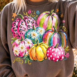 May include: A dark brown crewneck sweatshirt featuring a colorful graphic print of multiple pumpkins. The pumpkins are decorated with various patterns including leopard print, white polka dots, and white daisy flowers in shades of pink, yellow, and blue.