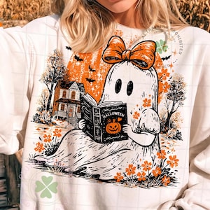 Op de afbeelding: Een wit sweatshirt met een afbeelding van een geest met een oranje strik, die een boek leest met de tekst Halloween en een pompoenillustratie. De sc&egrave;ne bevat een huis, bomen, vleermuizen en oranje bloemen tegen een grote oranje maan.