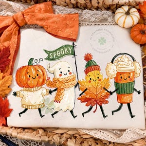 May include: A white t-shirt featuring a colorful illustration of a pumpkin, a ghost holding a green flag with the text SPOOKY, a maple leaf, and a coffee cup, all with smiling faces, surrounded by orange fabric, autumn leaves, and pumpkins.