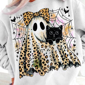 May include: A white sweatshirt featuring a graphic of a ghost wearing a leopard print bow and leopard print sheet. The ghost holds a black cat in front of a spider web background with small bats, stars, and hearts in black and orange ink.