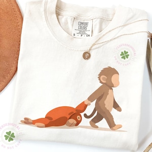 Puede incluir: Camiseta color crema con un mono de dibujos animados tirando de un animal rojo. La camiseta tiene una etiqueta Comfort Colors y un collar dorado. Un sombrero marrón y una planta seca están en el fondo.