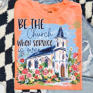 May include: Orange t-shirt with the text "BE THE Church WHEN SERVICE is OVER" and a watercolor painting of a church surrounded by flowers. The shirt is folded on top of a pair of blue jeans.