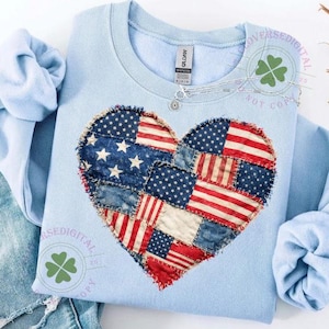 Puede incluir: Una sudadera azul claro con un diseño en forma de corazón hecho con parches de banderas americanas. La sudadera se combina con unos vaqueros de mezclilla azul claro con detalles desgastados. La imagen está tomada sobre una superficie blanca.