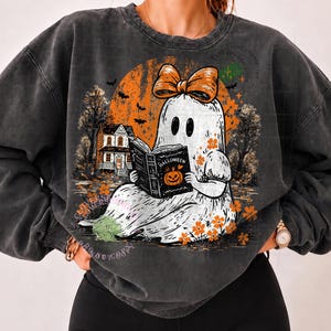 Op de afbeelding: Een antracietgrijze sweater met ronde hals en een afbeelding van een witte geest met een oranje strik, die een boek leest met de titel Halloween en een pompoenillustratie. De achtergrond toont een grote oranje maan, een huis en bomen.