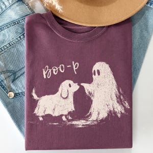 May include: A dark plum-colored crew neck t-shirt featuring a light-colored graphic print of a dog wearing a ghost costume standing next to a ghost. The text "Boo-p" is printed in a light-colored, handwritten-style font above the graphic.