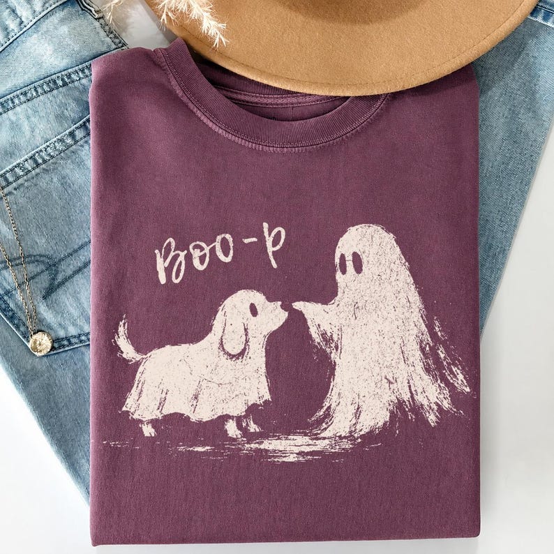 May include: A dark plum-colored crew neck t-shirt featuring a light-colored graphic print of a dog wearing a ghost costume standing next to a ghost. The text "Boo-p" is printed in a light-colored, handwritten-style font above the graphic.